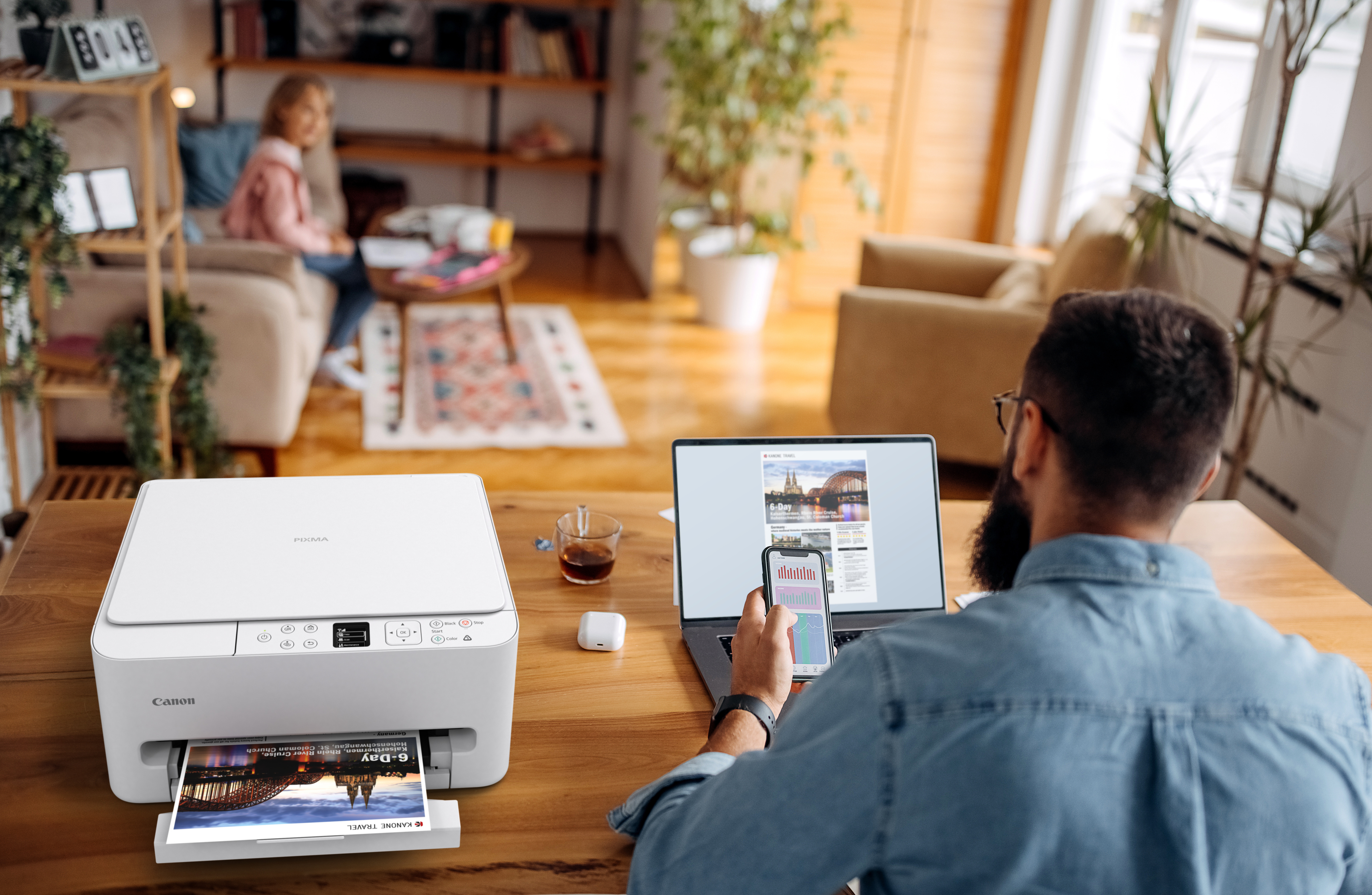 Ein Mann sitzt an einem Tisch mit einem Laptop, Drucker und Smartphone, im Hintergrund eine Frau auf einem Sofa.
