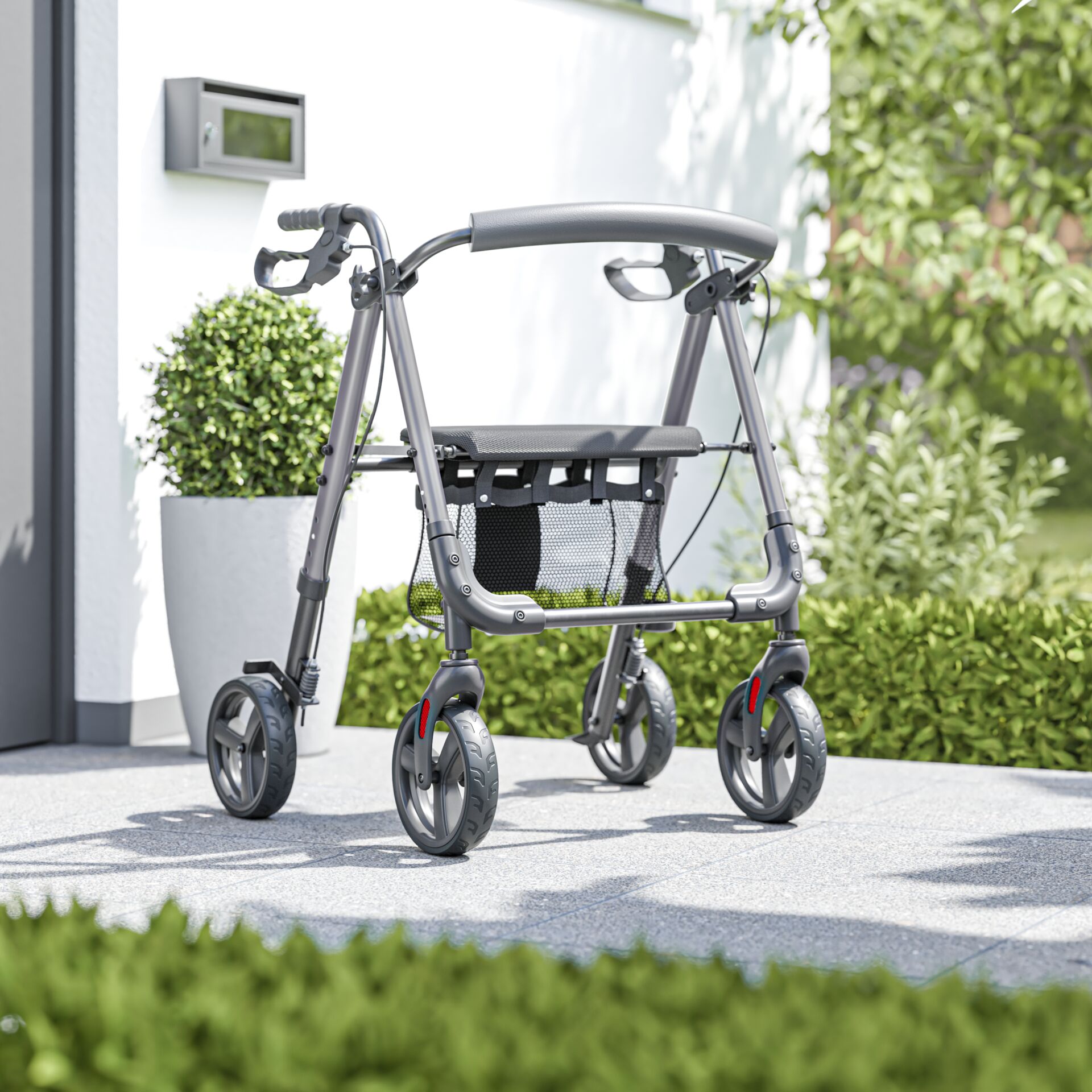 Silberner Rollator mit vier Rädern, abgestellt auf einer Terrasse vor einem weißen Gebäude mit Pflanzen im Hintergrund