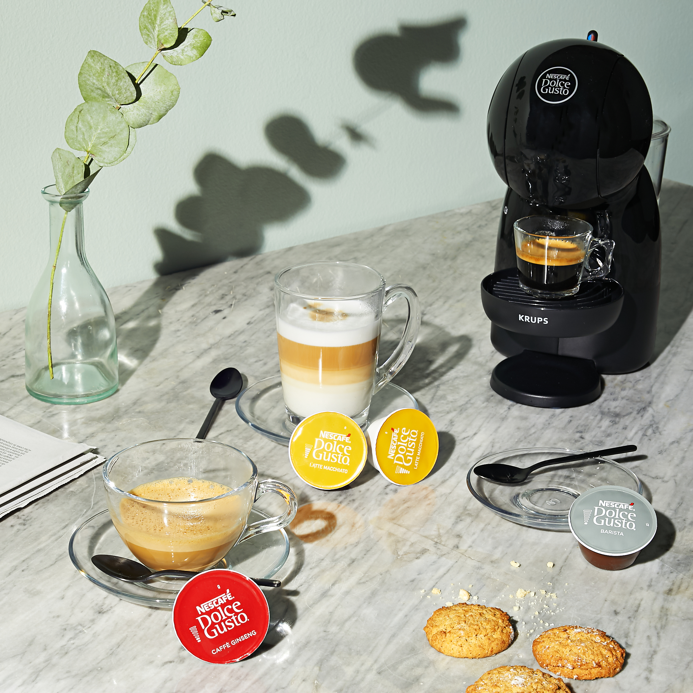 Schwarze Nescafé Dolce Gusto Kaffeemaschine, Cappuccino und Latte Macchiato in Glasbechern auf Marmortisch, umgeben von Kaffeekapseln und Keksen