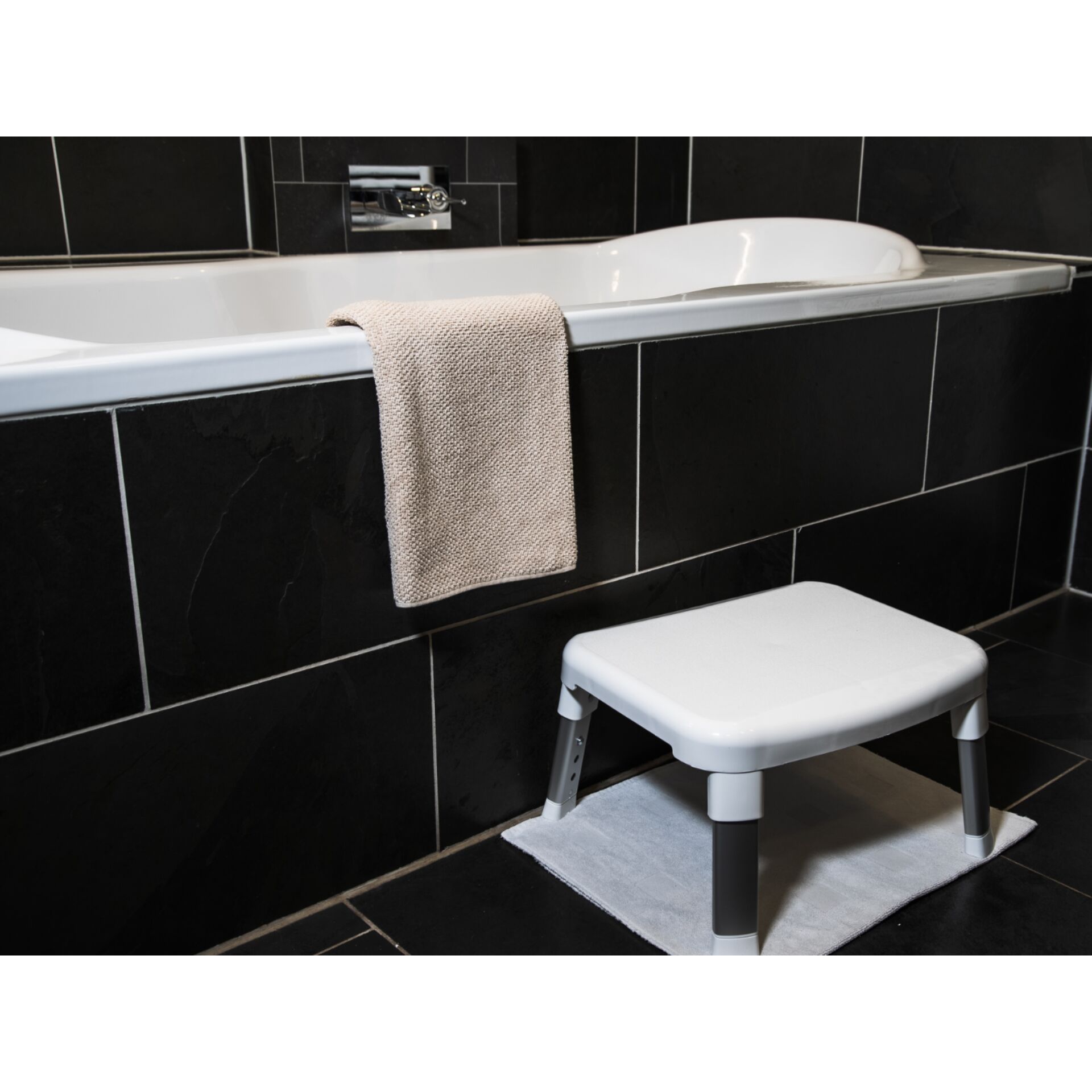 Ein weißer Hocker steht vor einer schwarz gefliesten Badewanne, auf der ein beiges Handtuch liegt.